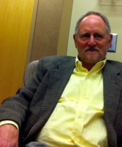 Portrait of Dr. Rutherford seated in an office chair, wearing a yellow button-down shirt and a gray blazer, with a neutral office background visible.