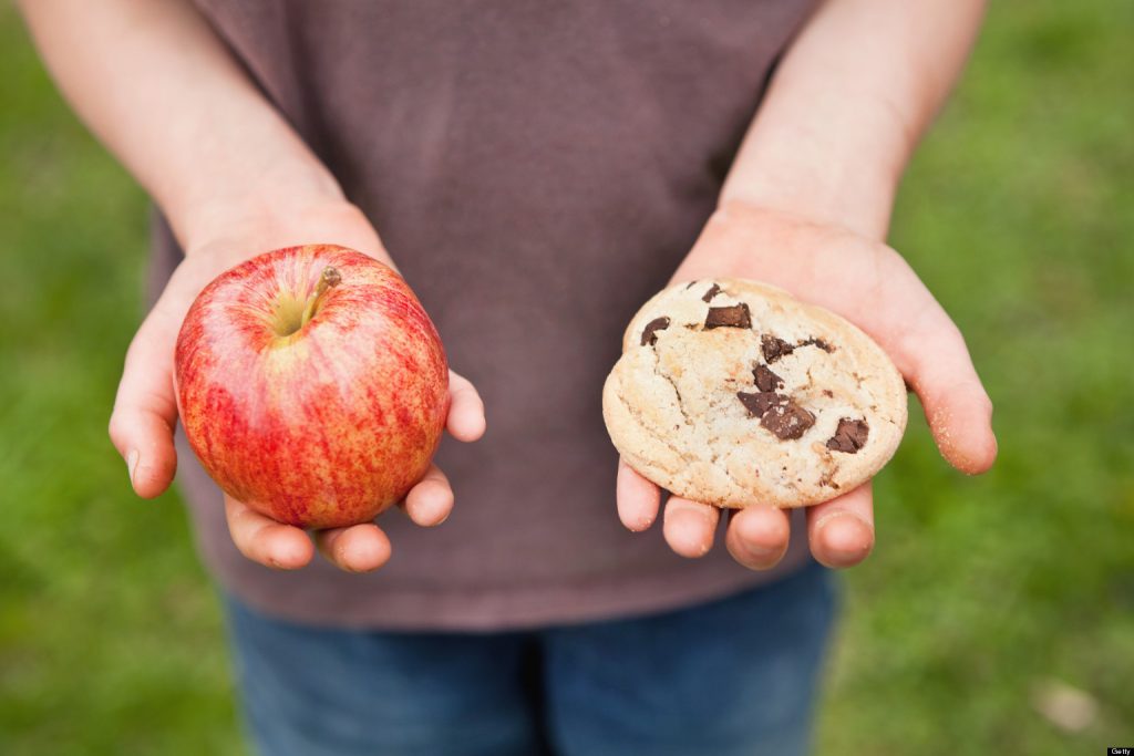 Child holding a red apple in one hand and a chocolate chip cookie in the other, illustrating a healthy food choice for kids.