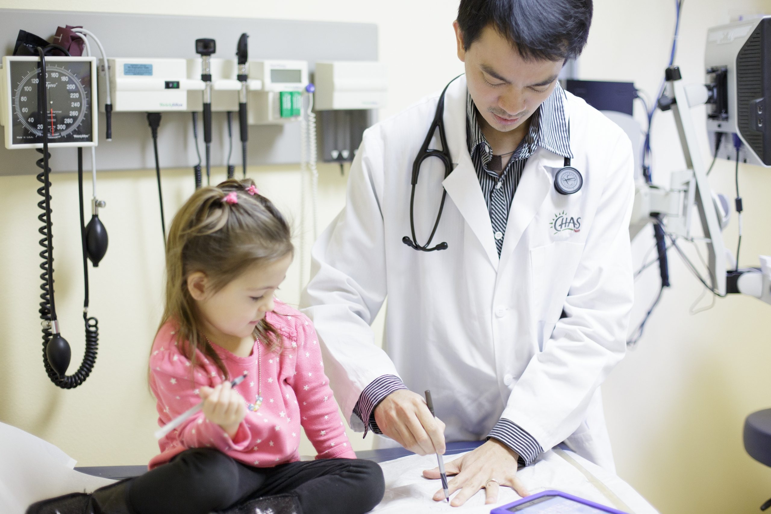 Healthcare provider at Valley Clinic reviews a clipboard with a child patient seated on an exam table in a medical exam room with diagnostic equipment on the wall.