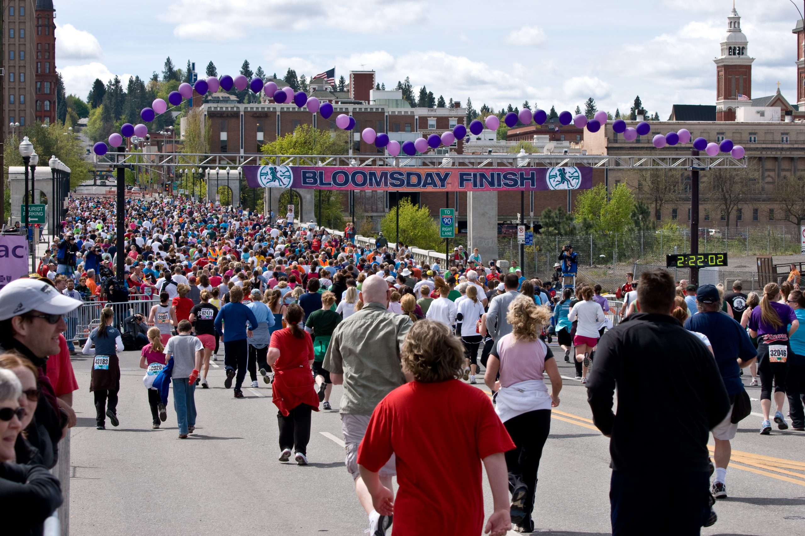 A large crowd of runners moves toward the finish line at the 2010 Bloomsday race in Spokane. A banner reading “BLOOMSDAY FINISH” stretches across the street beneath rows of purple balloons, with race volunteers, spectators, and surrounding city buildings visible in the background.
