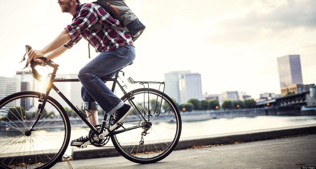 Person riding a bicycle along a city waterfront path during daylight hours, wearing casual clothing and a backpack, with urban buildings visible in the background, used to illustrate a blog post about biking to work.
