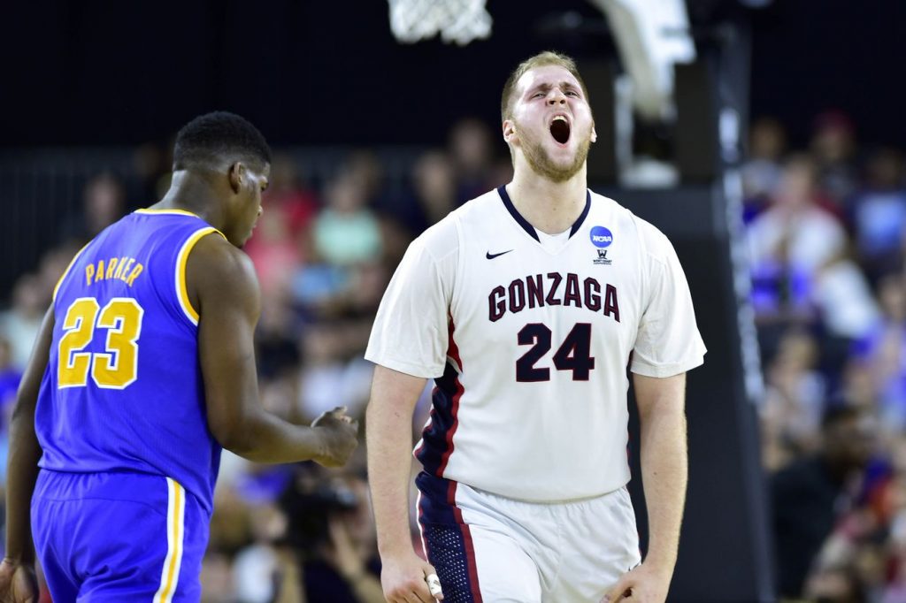 College basketball game scene from 2017. In the foreground, Przemek Karnowski wearing a white Gonzaga jersey with the number 24 stands with mouth open, arms at sides. To the left, another player in a purple jersey with yellow trim and the name ‘Parker’ and number 23 is turned partly away. A basketball hoop and blurred crowd are visible in the background.