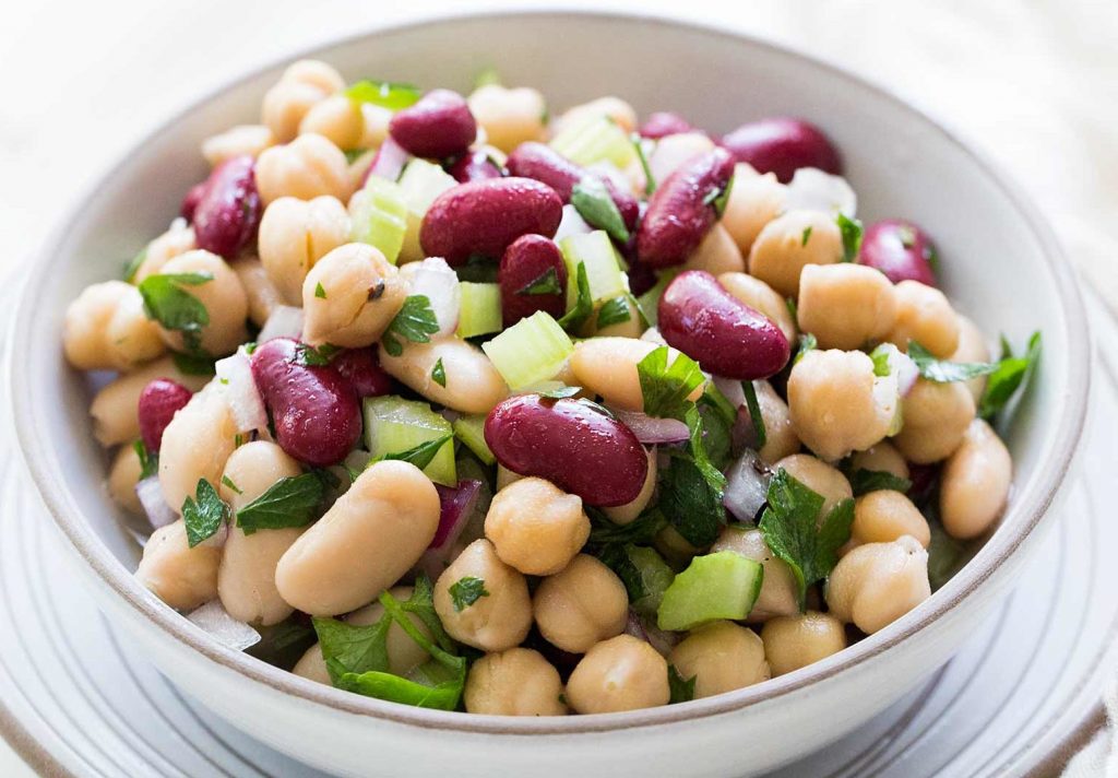 Bowl of three bean salad with kidney beans, chickpeas, and white beans mixed with chopped celery, red onion, and fresh herbs.