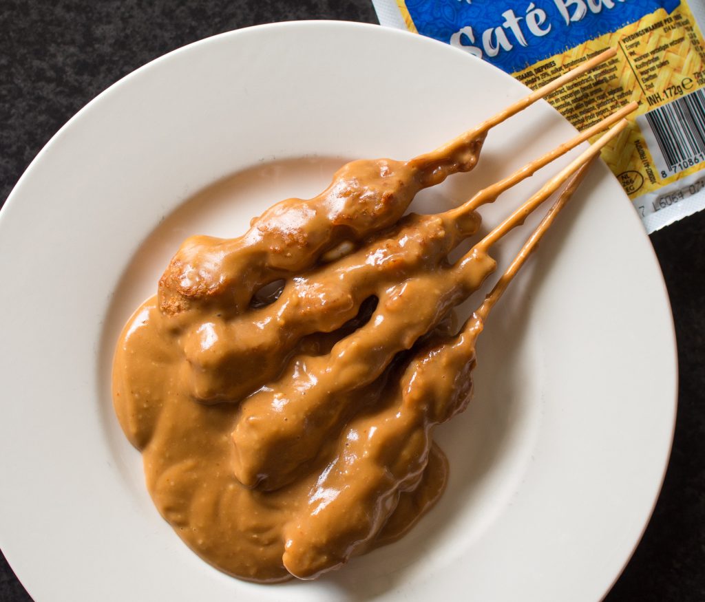 “Peanut dipping sauce pooled in a white bowl with skewers of satay coated in creamy peanut sauce, shown from above on a dark surface.