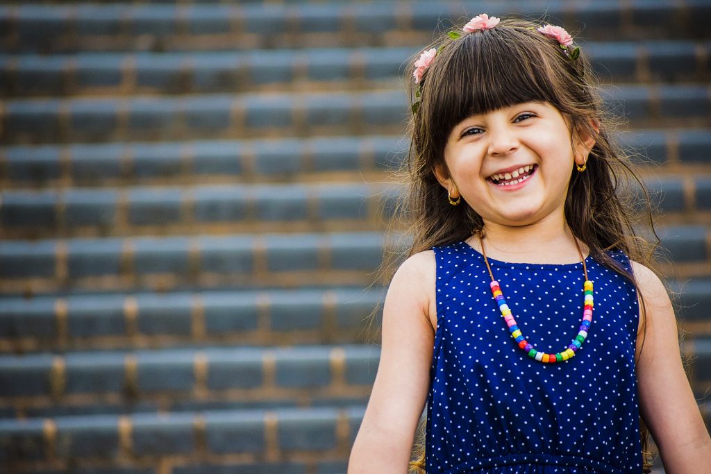 Child standing outdoors in front of wide brick steps, wearing a blue sleeveless dress with white polka dots and a colorful beaded necklace, with long dark hair and small flower accessories.