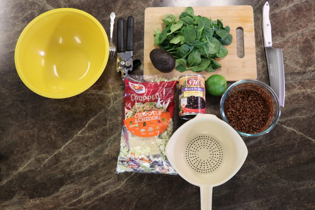 Ingredients for a power salad arranged on a countertop, including leafy greens, avocado, lime, black beans, cooked grains, chopped salad mix, and kitchen utensils ready for preparation.