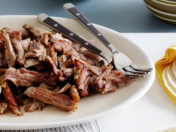 White plate filled with shredded pork carnitas with crispy, browned edges, two metal forks resting on the plate, placed on a white surface beside a folded yellow napkin.