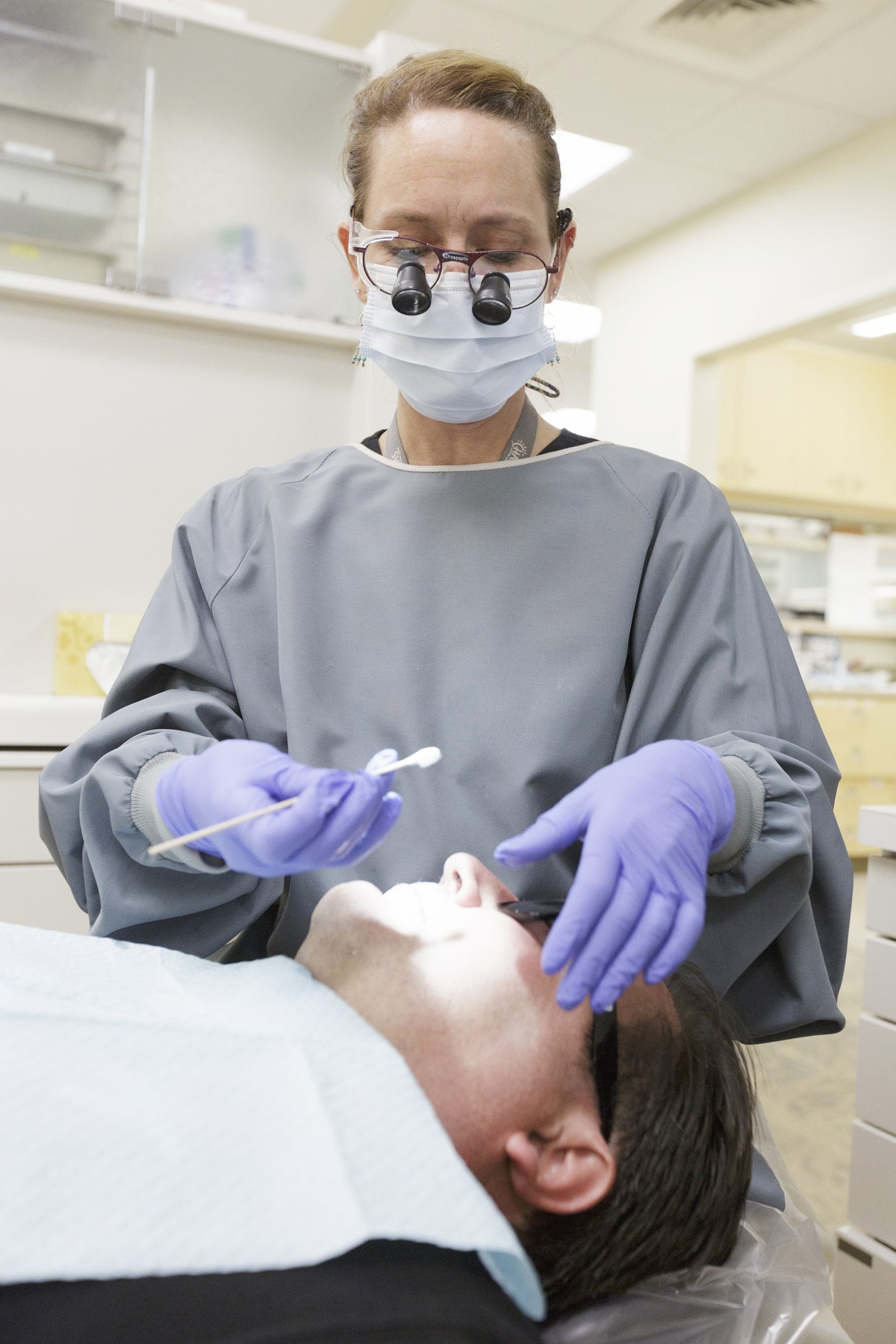 CHAS Health provider wearing a mask and gloves examines a patient lying in a dental chair under an overhead exam light.