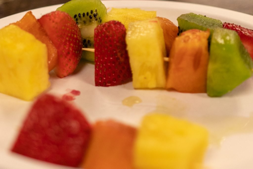Close-up of festive fruit kabobs arranged on a white plate, featuring alternating pieces of strawberries, pineapple, kiwi, and melon threaded onto wooden skewers.