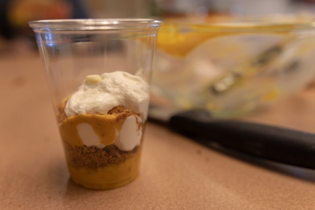 Pumpkin pie parfait served in a clear plastic cup, layered with pumpkin filling, whipped cream, and graham cracker crumbs on a kitchen counter.