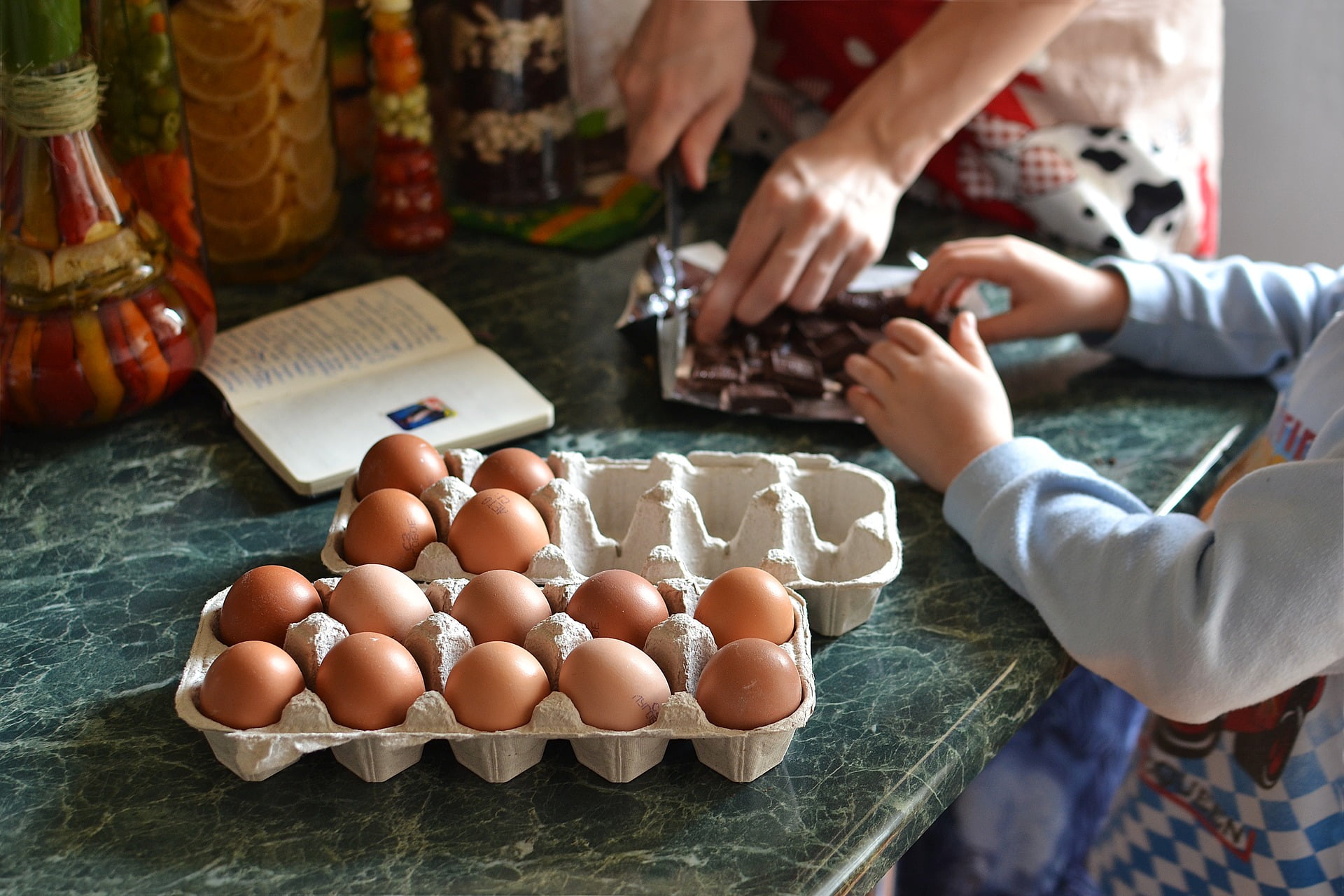 A carton of brown eggs sits on a green marble countertop next to an open cookbook. Two people work together nearby, with one set of hands chopping chocolate and another set of hands reaching toward the pieces.