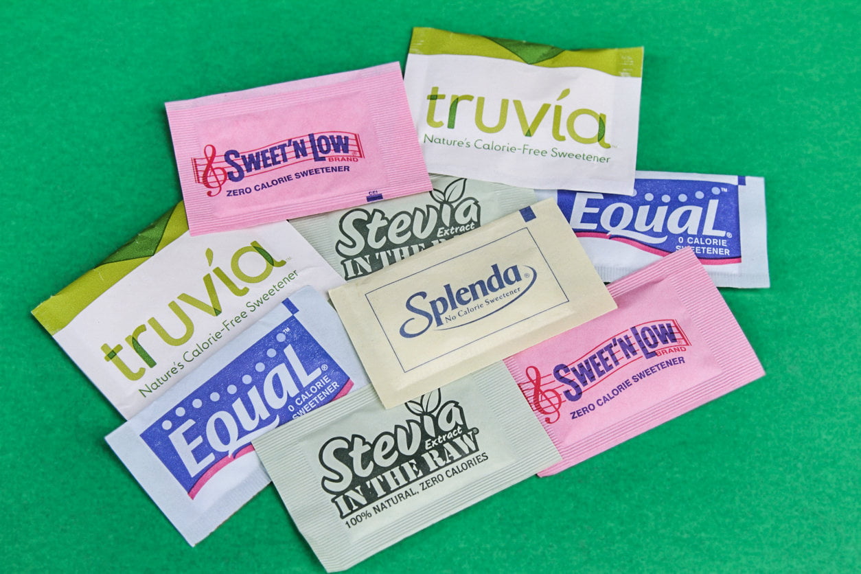 A variety of single‑serve sweetener packets are spread out on a green surface, including brands such as Truvia, Sweet’N Low, Equal, Splenda, and Stevia In The Raw.