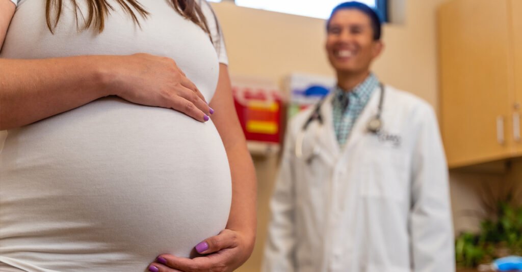 Close-up of a pregnant patient’s abdomen with hands resting on the belly, with a healthcare provider in a white coat standing in the background of a clinic exam room.