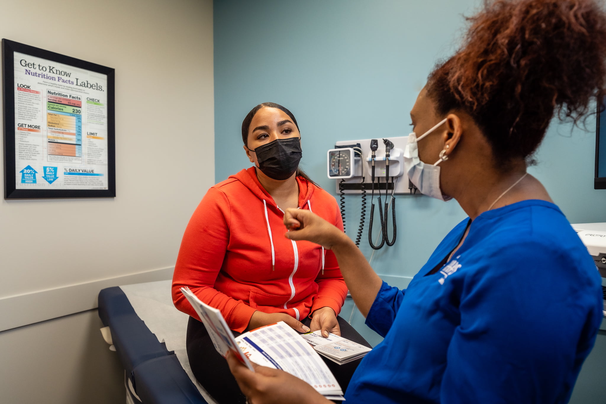 A healthcare worker in blue scrubs speaks with a patient seated on an exam table in a medical room. The worker holds printed educational materials while gesturing toward the patient. Medical equipment, including a wall‑mounted blood pressure monitor and otoscope, is visible in the background along with a framed nutrition facts poster.