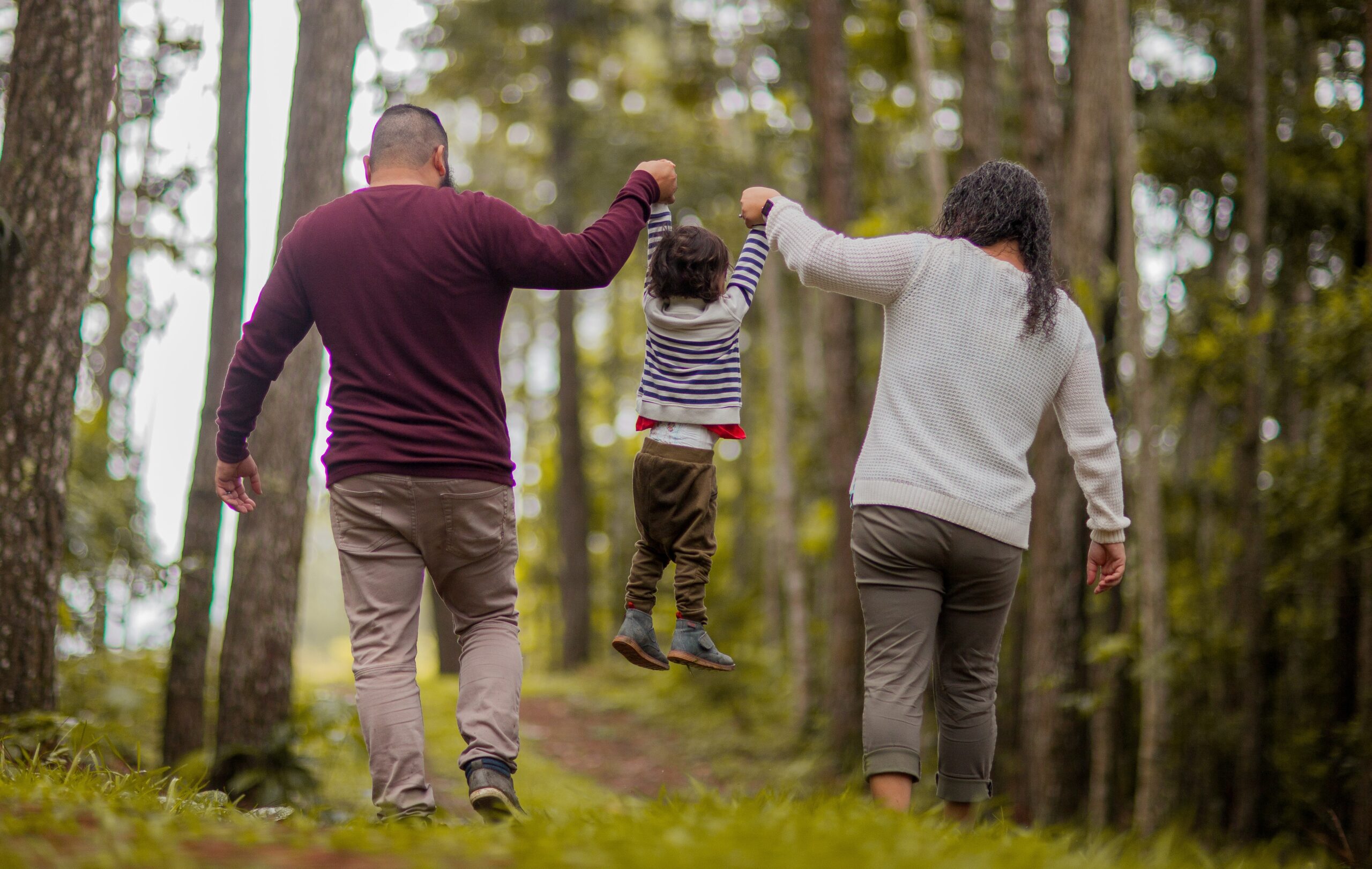 Three people walk along a forest path, with the two adults each holding one of the child’s hands and lifting the child slightly off the ground as they move forward. Tall trees and soft natural light surround them.