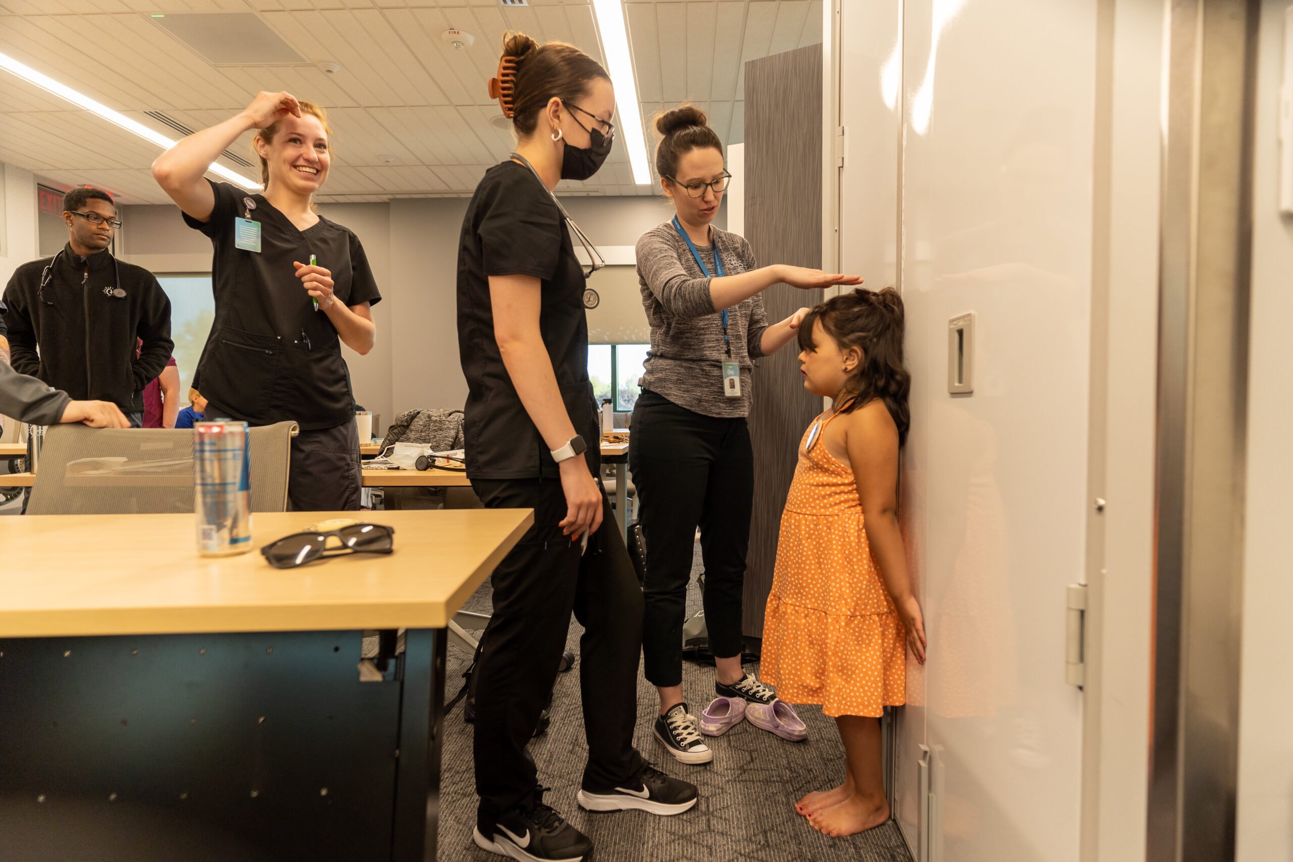 Medical assistant apprenticeship students practice taking a child’s height measurement in a classroom setting. One student uses a wall‑mounted measuring tool while another observes, and additional students stand nearby. A child stands barefoot against the measurement wall, and desks, chairs, and classroom equipment are visible in the background.