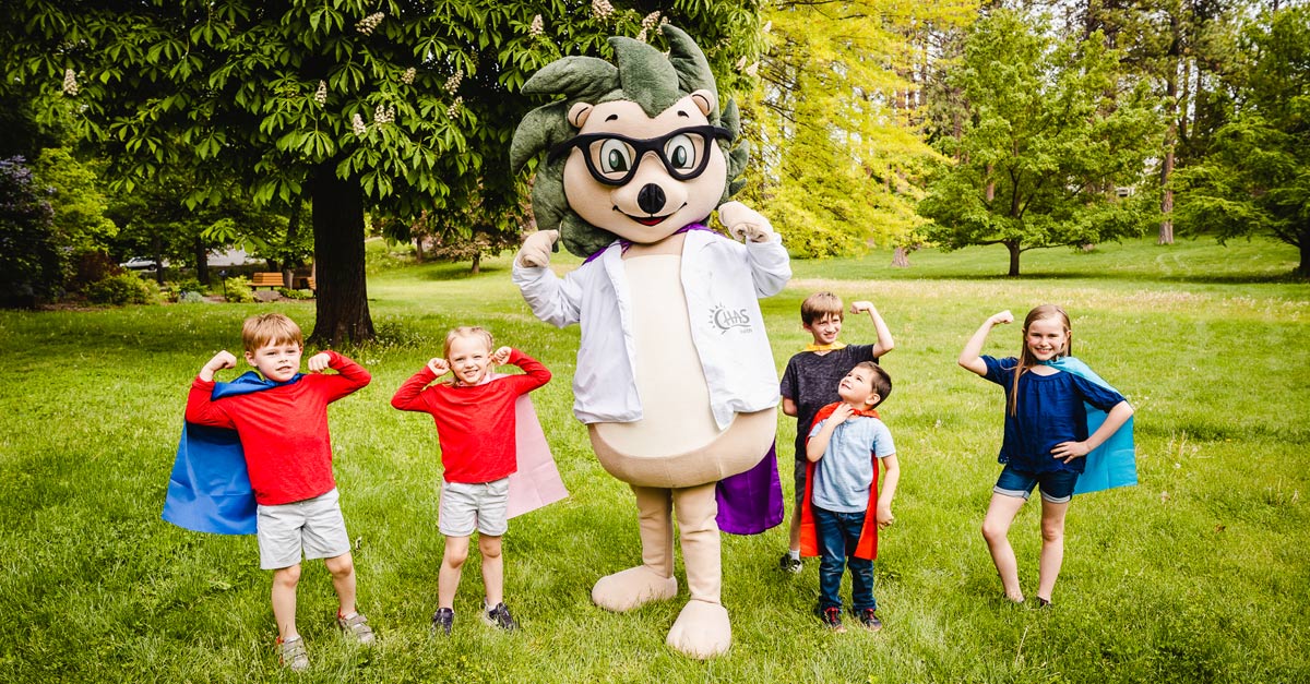 CHAS Health mascot Chazzy stands in a grassy park with several children wearing capes, all posing with flexed arms.
