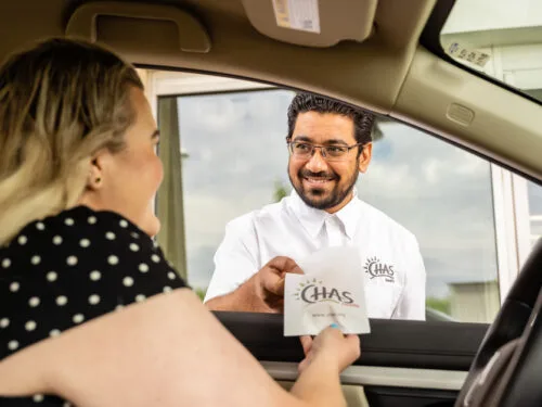 A patient in a vehicle receives a CHAS Health pharmacy bag from a staff member standing outside the driver-side window at a drive‑up service area.