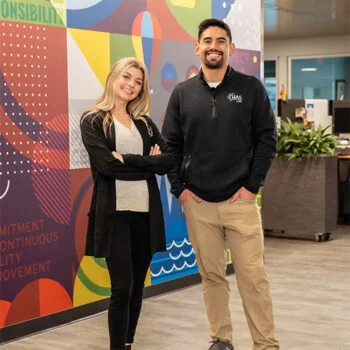 Two CHAS Health employees stand in a brightly decorated office space featuring a colorful wall mural. They appear confident and professional, representing the welcoming workplace environment promoted in CHAS Health career opportunities.