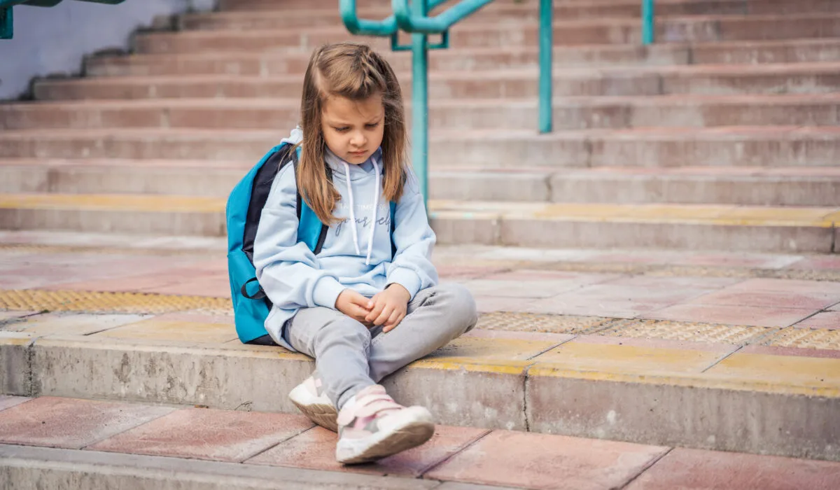 A child wearing a light‑colored hoodie, gray pants, and sneakers sits on outdoor steps with a backpack resting behind them. The wide concrete staircase and metal railings extend upward in the background.