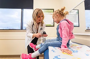 A healthcare professional kneels beside a young child seated on an exam table in a medical clinic, holding a medical instrument while interacting with the child.