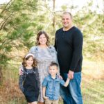 Portrait of the Proett family smiling outdoors, with family members sitting closely together in fall scenery.
