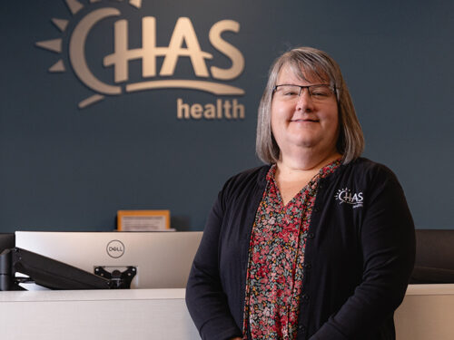 A patient resource counselor stands at a reception desk inside a CHAS Health clinic. The individual is wearing a black cardigan with the CHAS Health logo over a patterned top.