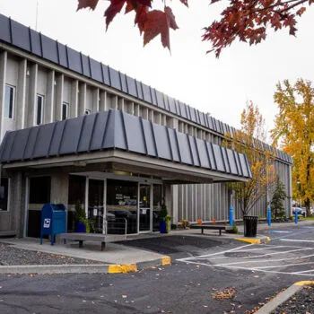 Exterior view of the CHAS Health Lidgerwood Clinic building, showing the front entrance with a covered awning, benches near the entrance, a parking area in the foreground, and autumn trees with fall-colored leaves surrounding the building.
