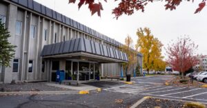 Exterior view of the CHAS Health Lidgerwood Clinic building, showing the front entrance with a covered awning, benches near the entrance, a parking area in the foreground, and autumn trees with fall-colored leaves surrounding the building.
