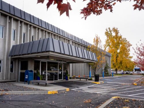 Exterior view of the CHAS Health Lidgerwood Clinic building, showing the front entrance with a covered awning, benches near the entrance, a parking area in the foreground, and autumn trees with fall-colored leaves surrounding the building.