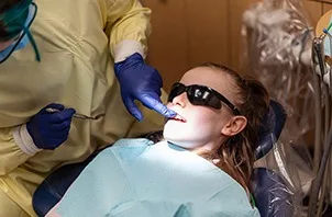 Patient reclining in a dental chair wearing protective eyewear while a dental provider wearing gloves and a mask performs an oral exam under bright exam lighting.