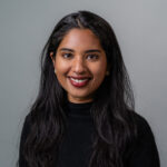 Dr. Sera Jacob standing against a plain background with long dark hair and wearing a dark top.