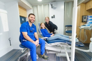 Child lying in a dental exam chair inside a mobile dental clinic, with clinic staff seated and standing nearby.