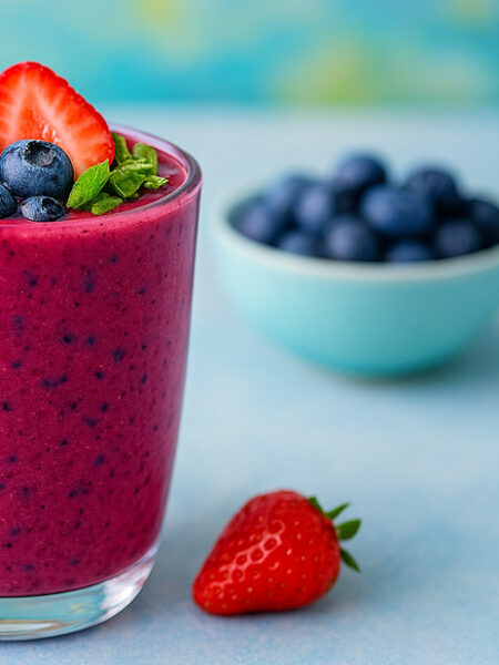 Colorful berry smoothie in a glass topped with strawberries, blueberries, and mint, with blurred fruit in the background.