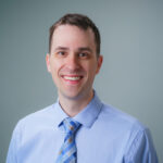 Professional head‑and‑shoulders portrait of an individual wearing a light blue dress shirt and striped tie, photographed against a soft, neutral background with studio lighting.