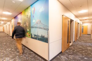 Spokane Pediatrics clinic hallway with exam room doors along a carpeted corridor, featuring a large colorful mural on the wall depicting local scenery and landmarks, illuminated by bright overhead lighting in a clean, modern healthcare setting.