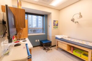 Spokane Pediatrics exam room with a clean, child‑friendly layout, featuring an exam table with colorful paper, wall‑mounted medical equipment, a sink and workstation, storage bins for toys or supplies, and a window providing natural light.