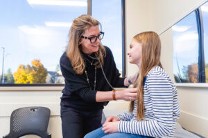 Pediatric provider at Spokane Pediatrics using a stethoscope to examine a child seated in an exam room, with large windows providing natural light and a clean, welcoming clinical setting.