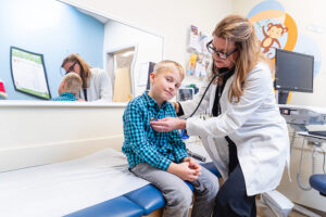 Pediatric healthcare provider examining a child seated on an exam table at Spokane Pediatrics, using a stethoscope in a bright, child‑friendly exam room with medical equipment, a wall mirror, and colorful decor visible in the background.
