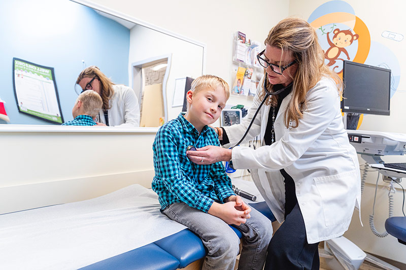 Nhân viên y tế nhi khoa đang khám cho một đứa trẻ ngồi trên bàn khám tại phòng khám nhi Spokane Pediatrics, sử dụng ống nghe trong một phòng khám sáng sủa, thân thiện với trẻ em, với các thiết bị y tế, gương treo tường và đồ trang trí đầy màu sắc ở phía sau.