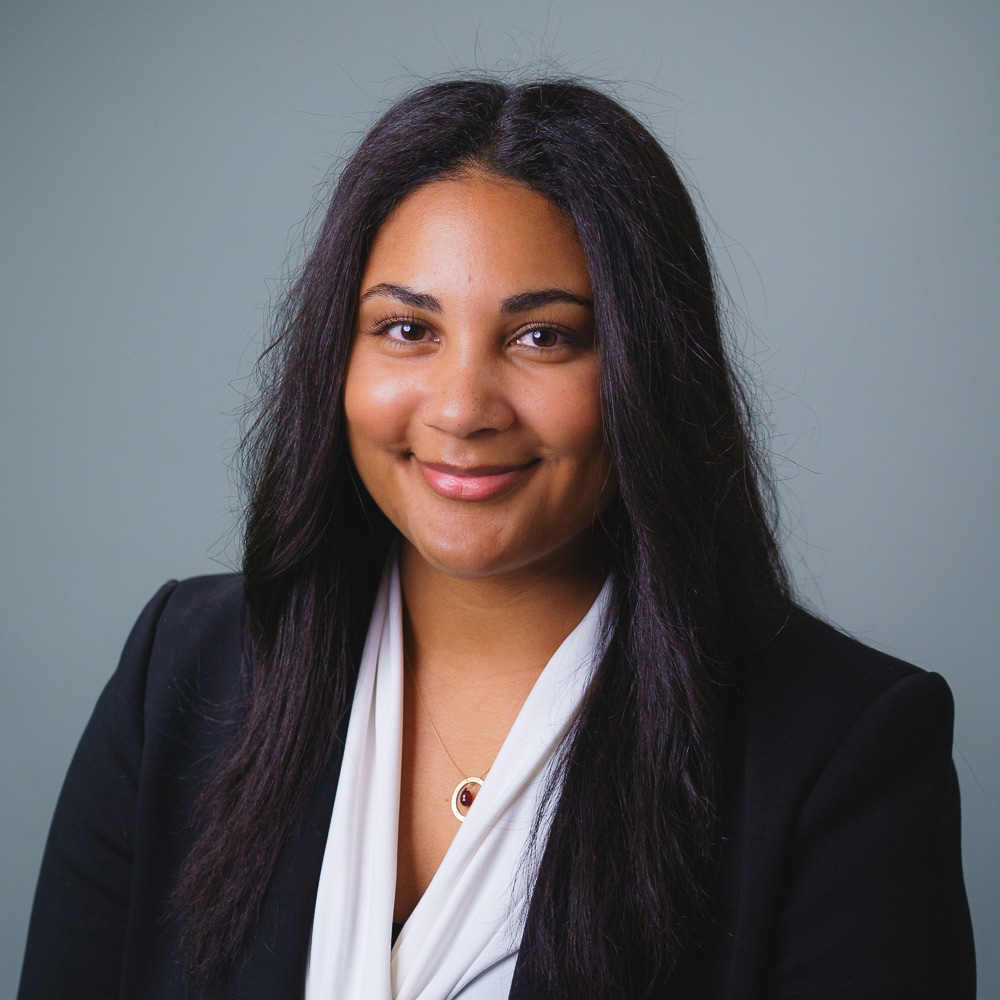 Professional headshot of a person with long dark hair, wearing a black blazer over a white blouse, facing forward against a neutral gray background.