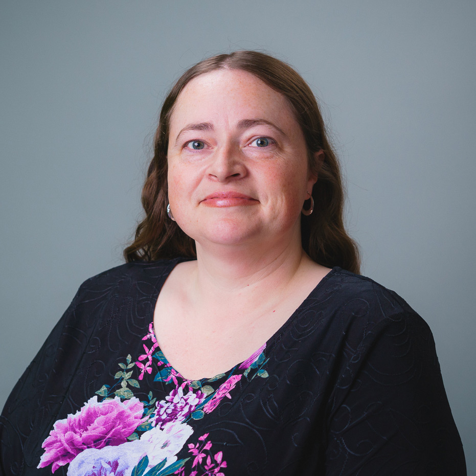 Professional headshot of a person with shoulder-length hair wearing a black top with a colorful floral pattern, against a gray background.