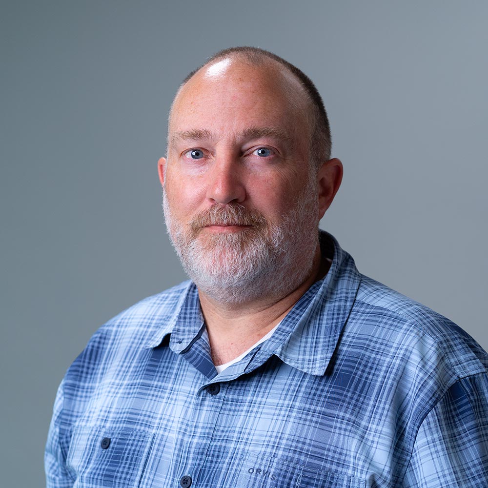 Head-and-shoulders portrait of an adult with a short haircut and a full beard, wearing a blue plaid shirt, against a plain gray background.