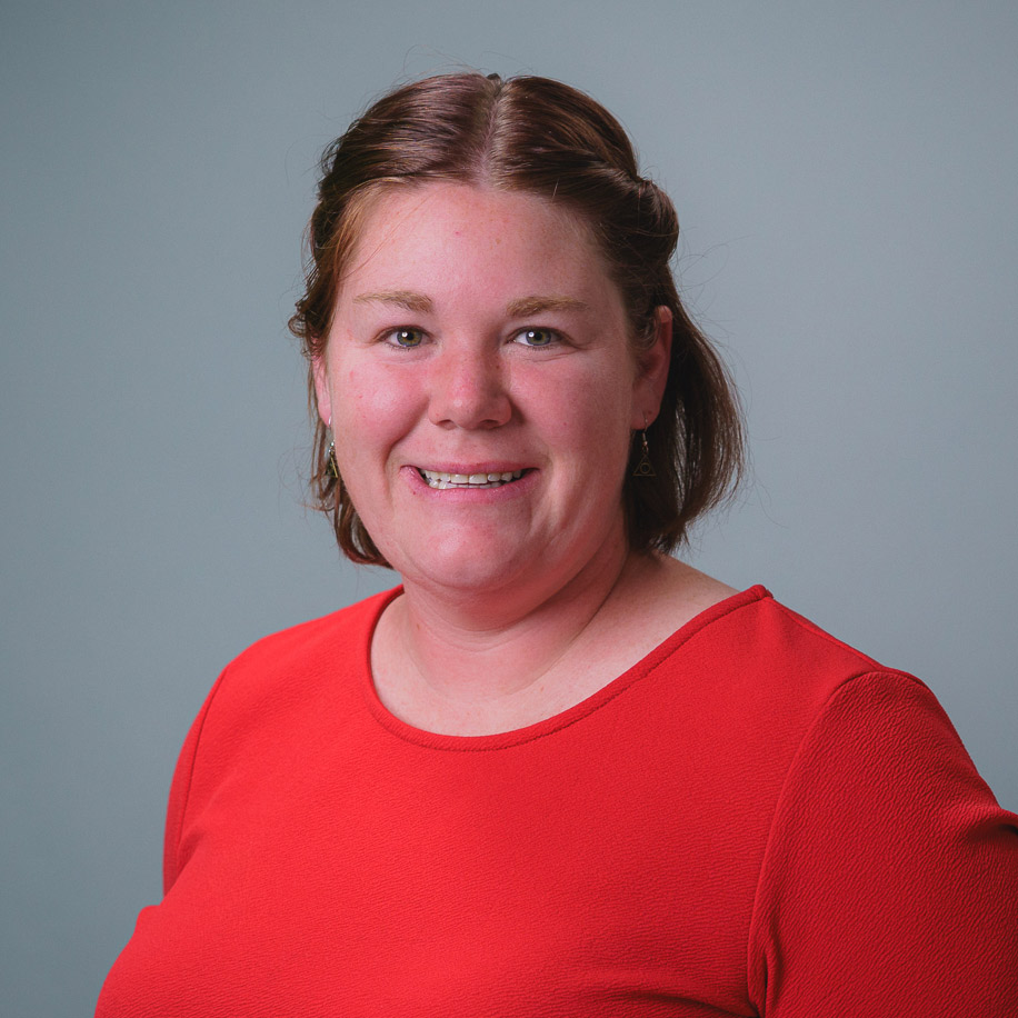 Headshot of a person wearing a bright red top against a plain gray background, facing forward with hair pulled back and a slight smile.