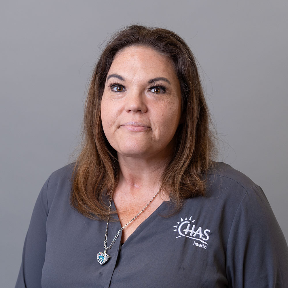 Portrait headshot of a person wearing a gray CHAS Health shirt and a silver pendant necklace, posed against a light gray background.