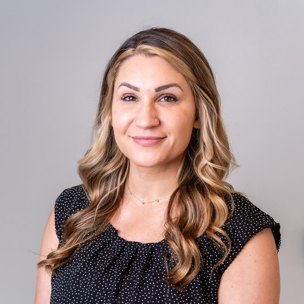 Professional headshot of a person with long wavy highlighted hair wearing a black patterned blouse against a light gray background.