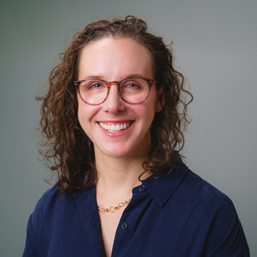 Headshot of a person with curly hair and glasses wearing a navy blue shirt against a gray background.