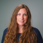 Professional headshot of a person with long brown hair wearing a navy blue top against a gray background.