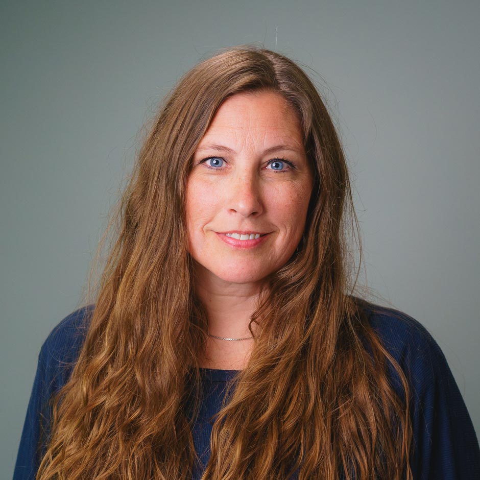 Professional headshot of a person with long brown hair wearing a navy blue top against a gray background.