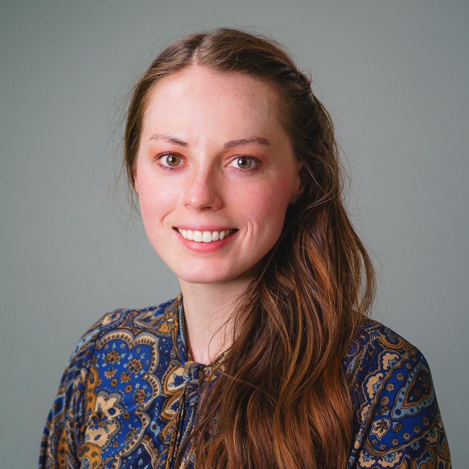 Professional headshot of a person with long brown hair wearing a blue patterned blouse against a gray background.